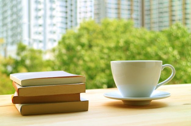 Pilha de livros e uma xícara de bebidas quentes na mesa perto da janela com folhagem verde e alta construção