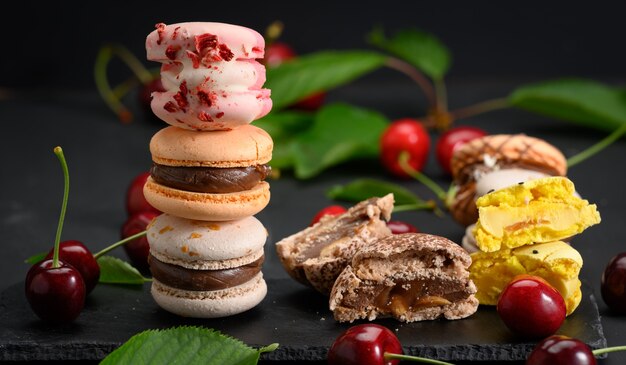 Pilha de macarons multicoloridos e cerejas vermelhas maduras em fundo preto de madeira, close-up