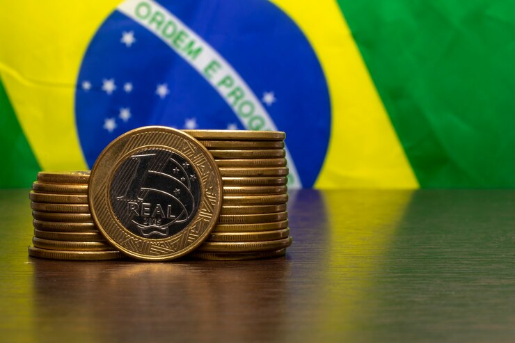 Pilha de moedas brasileiras com um real em destaque e a bandeira do Brasil ao fundo