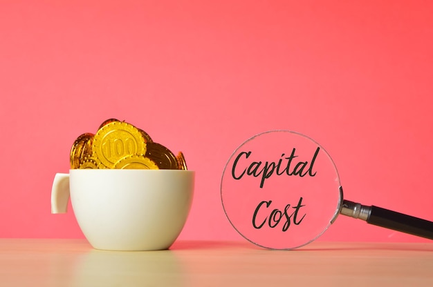 Pilha de moedas em uma caneca e lupa com a frase CAPITAL COST