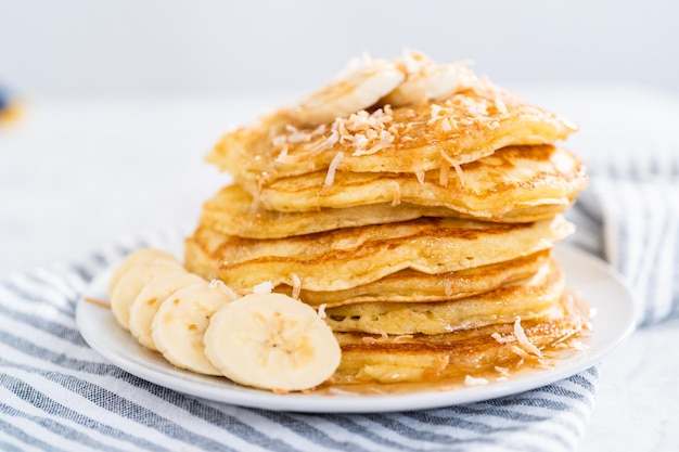 Pilha de panquecas de banana de coco recém-assadas, guarnecidas com bananas fatiadas, coco torrado e xarope de bordo