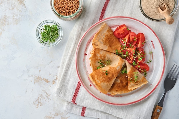 Pilha de panquecas de crepes de farinha de trigo sarraceno sem glúten com tomate cereja e microgreens de rúcula na chapa branca, cozimento caseiro saudável no café da manhã. Brincar. Copie o espaço.