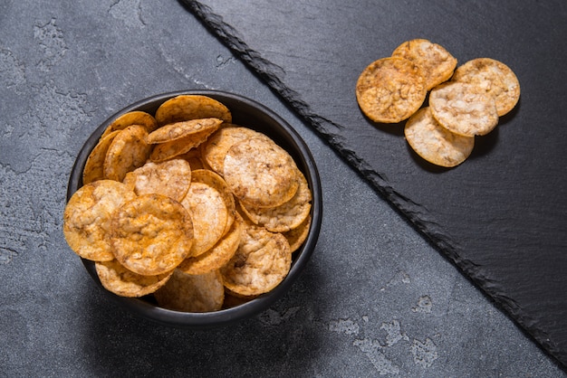 Pilha de vista superior de chips de arroz orgânico, crocante, assado, grãos integrais com especiarias. lanche saudável sem glúten. bacia cerâmica preta sobre fundo de pedra cinza