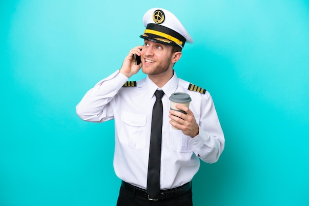 Piloto de avião caucasiano isolado em um fundo azul segurando café para levar e um celular