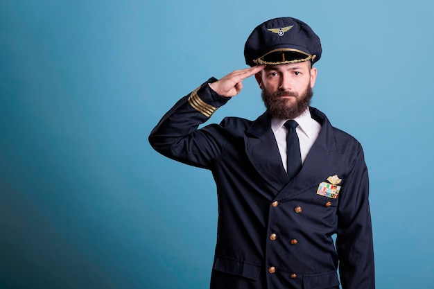 Piloto de avião sério saudando, vestindo uniforme e retrato de vista frontal de chapéu, capitão de avião olhando para a câmera. Aviador da academia de aviação com distintivo de asas de companhia aérea na jaqueta