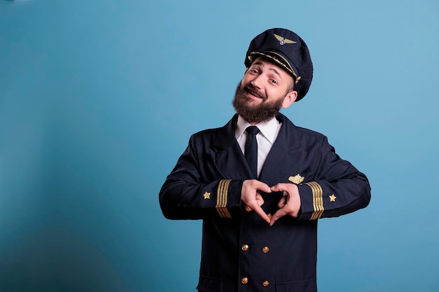 Piloto de avião sorridente mostrando símbolo de amor em forma de coração com dedos, gesto de romance conceitual. Capitão de aeronave amigável em uniforme expressando sentimentos de afeto vista frontal plano médio