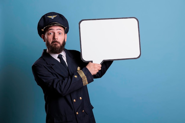 Piloto de avião surpreso segurando balão branco vazio com espaço de cópia, aviador com simulação de quadro de comunicação. Capitão de avião de uniforme em pé com balão de mensagem em branco