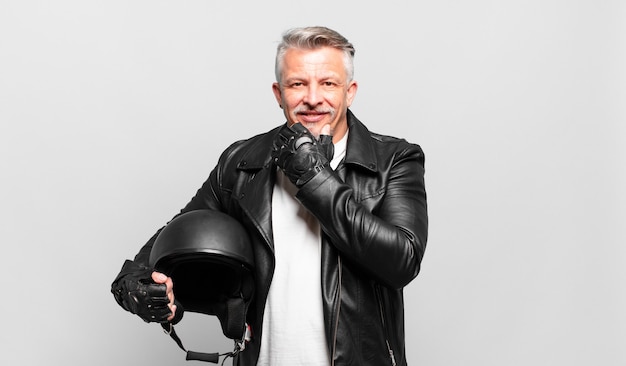 Piloto de motocicleta sênior sorrindo com uma expressão feliz e confiante com a mão no queixo, pensando e olhando para o lado