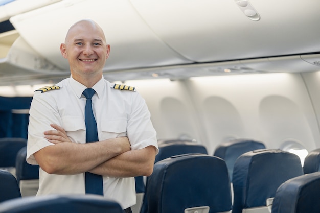 Piloto masculino alegre en uniforme manteniendo los brazos cruzados y sonriendo a la cámara mientras está de pie dentro del avión. Transporte, concepto de tripulación aérea