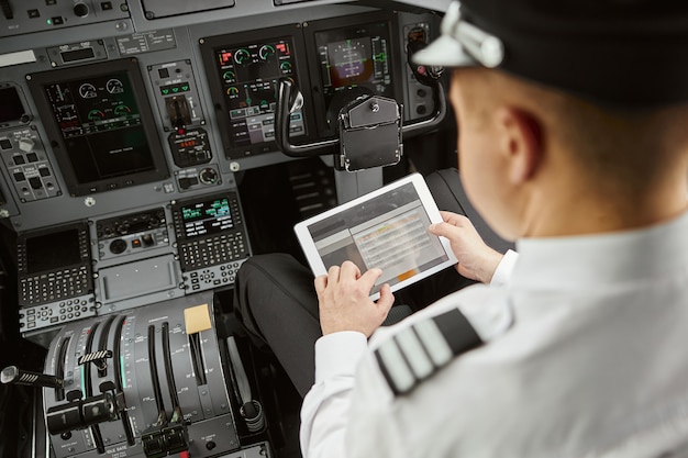 Piloto masculino usa tablet digital no jato do avião de passageiros. Interior do cockpit em avião moderno com painel e navegação aérea. Vista superior do uniforme de desgaste do homem. Aviação Civil. Conceito de viagem aérea