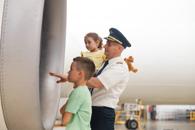 Piloto en uniforme mostrando partes del avión a dos niños pequeños que vinieron de excursión al hangar de aviones. Aviones, excursiones, concepto de infancia.