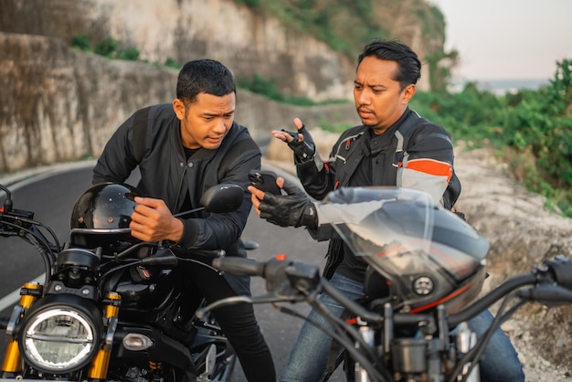 Pilotos asiáticos sentados em motos e conversando seriamente enquanto seguram o telefone