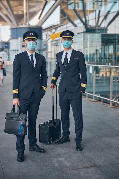 Pilotos jóvenes guapos con máscaras protectoras sosteniendo bolsas de viaje y mirando a la cámara mientras esperan el vuelo en el aeropuerto