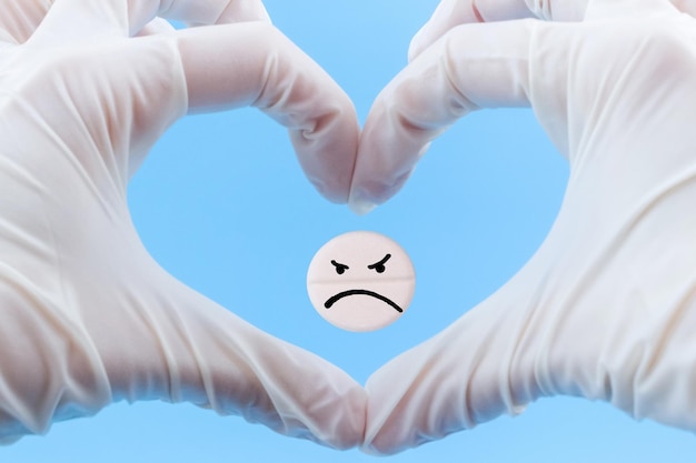 Pílula com um emoticon triste no coração das mãos em luvas médicas em um fundo azul cuidados de saúde