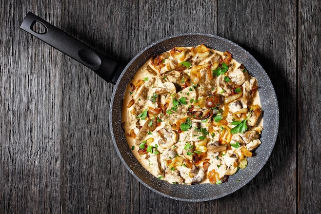 Pilz- und Kalbsleber-Stroganoff in einer Pfanne auf einem dunklen Eichenholztisch, flache Lage, amerikanische Küche