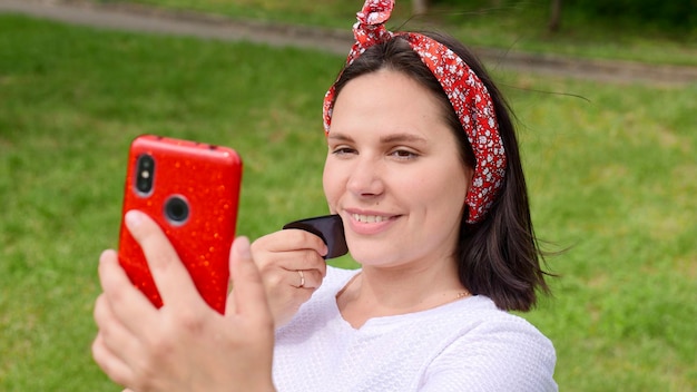 Pin Up Una mujer morena de unos 30 años sonríe y se masajea la cara con un raspador de gouache Gua Sha afuera frente al teléfono Masaje facial de gouache de entrenamiento en línea