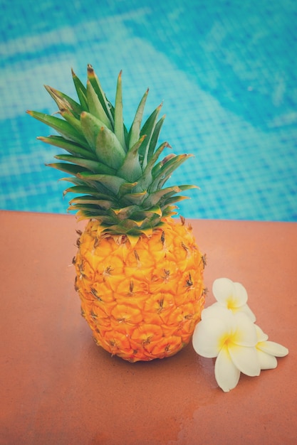 Piña amarilla entera cruda con flores y piscina de azulejos, tonos retro