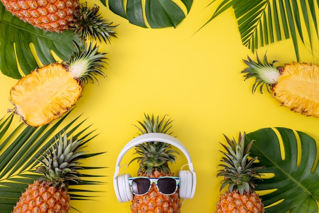 Piña divertida con auriculares blancos, escuchar música, aislado sobre fondo amarillo con hojas de palmeras tropicales, vista superior, concepto de diseño plano.