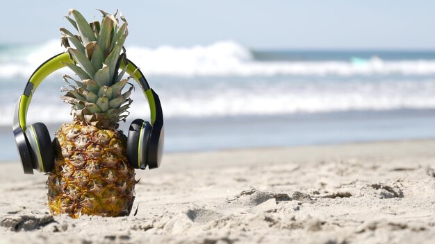 Piña divertida en auriculares, playa de arena, olas de agua de mar azul, costa del Pacífico de California, Estados Unidos. Fruta exótica de verano tropical disfrutando de las vacaciones y la música en el paraíso. Ananas tomando el sol en la orilla