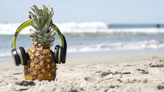 Piña divertida en auriculares, playa de arena, olas de agua de mar azul, costa del Pacífico de California, Estados Unidos. Fruta exótica de verano tropical disfrutando de las vacaciones y la música en el paraíso. Ananas tomando el sol en la orilla