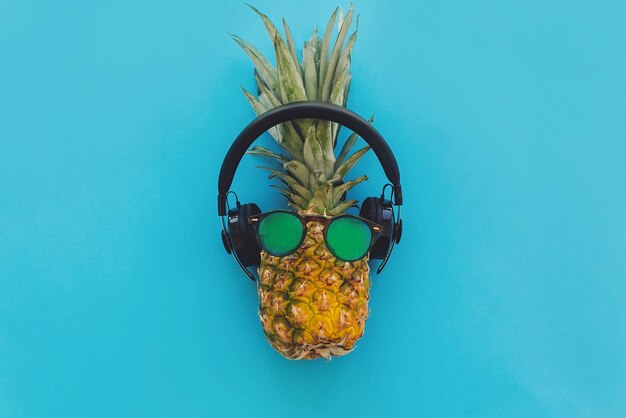 Foto piña en elegantes gafas de sol hipster y auriculares negros sobre fondo de papel de moda azul plano moderno concepto de vacaciones de verano fruta plana fiesta en la piscina y tema musical