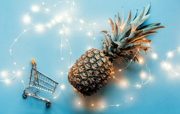 Piña fresca con luces de hadas y carrito de compras sobre fondo azul.