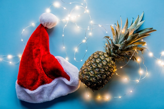 Piña fresca con luces de hadas y sombrero de Navidad sobre fondo azul.