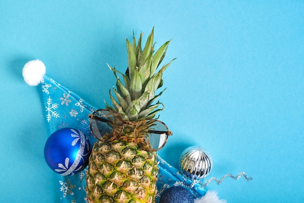 Piña en santa sombrero y gafas de sol, decoración navideña sobre fondo azul, vacaciones de invierno en concepto de trópicos