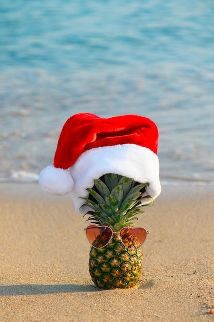 Una piña en el sombrero de Santa Claus con gafas de sol en forma de corazón en un fondo de Navidad de arena