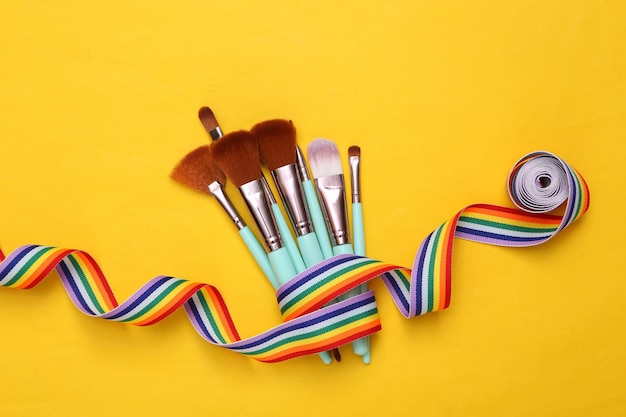 Pinceles de maquillaje con cinta de arco iris sobre fondo amarillo Diseño de belleza