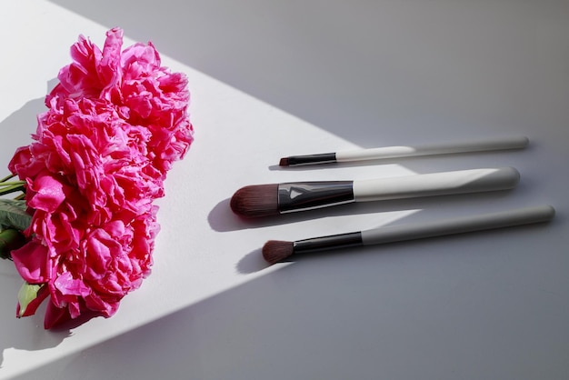 Foto pinceles de maquillaje y peonías rosas sobre un fondo blanco.