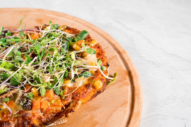 Pinsa romana vegana con champiñones, espárragos, maíz, guisantes, zanahorias, queso y salsa, aderezado con microgreens sobre tabla de madera.