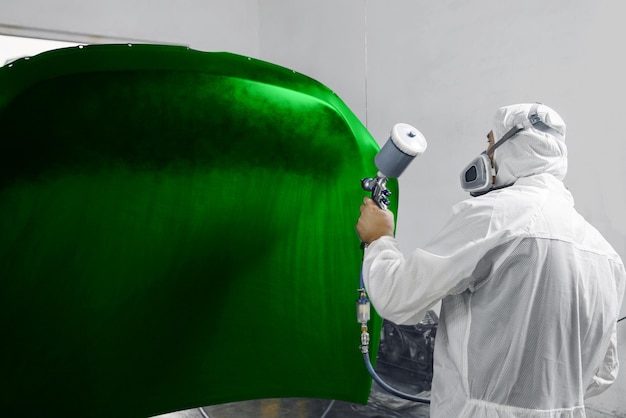 Pintador de reparación de automóviles con máscara y ropa de trabajo protectora pintando el capó del automóvil a color verde