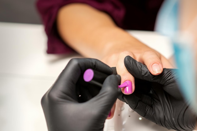 Pintar as unhas de uma mulher. Mãos de manicure em luvas pretas, aplicando esmalte rosa nas unhas femininas em um salão de beleza.