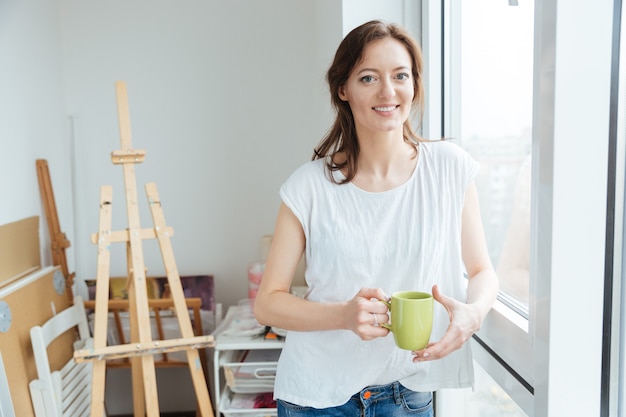 Pintor alegre hermosa mujer bebiendo té cerca de la ventana en el estudio de arte