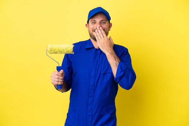 Pintor brasileiro isolado em fundo amarelo feliz e sorridente cobrindo a boca com a mão