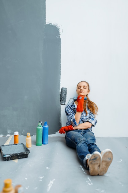 Pintor de casas femenino con rodillo de pintura sentado en el suelo después del trabajo. Reparación de viviendas, mujer feliz haciendo renovación de apartamentos