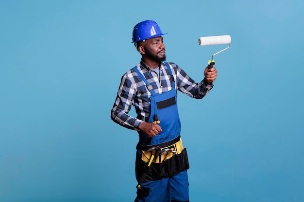 Pintor de casa fazendo reforma de interiores, pintando paredes com rolo. Trabalhador da construção civil vestindo uniforme e capacete protetor isolado sobre fundo azul, studio um tiro.