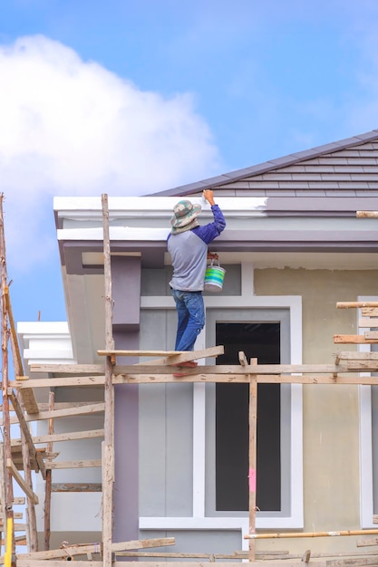 Pintor em andaimes de madeira está pintando beirais de telhado da estrutura moderna da casa no canteiro de obras