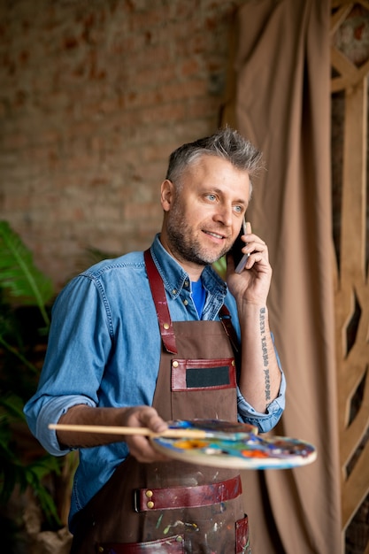 Pintor exitoso de mediana edad con paleta y pincel consultando con su maestro en el teléfono móvil en el estudio