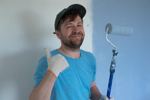 pintor con gorra de camiseta azul y guantes pintando una pared con un rodillo de pintura mostrando el pulgar hacia arriba