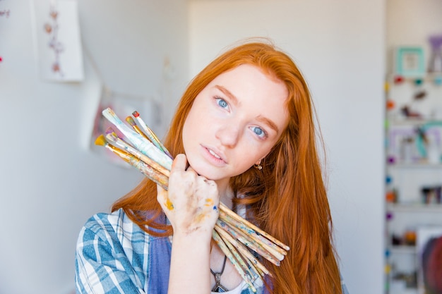 Pintor joven pensativo hermoso con el pelo rojo largo que sostiene los pinceles en el estudio de arte