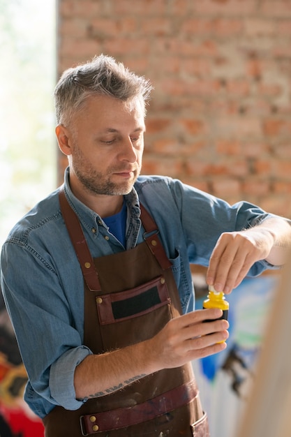 Pintor maduro en ropa de trabajo destapando un tarro de plástico con gouache antes de trabajar sobre una nueva pintura en el estudio
