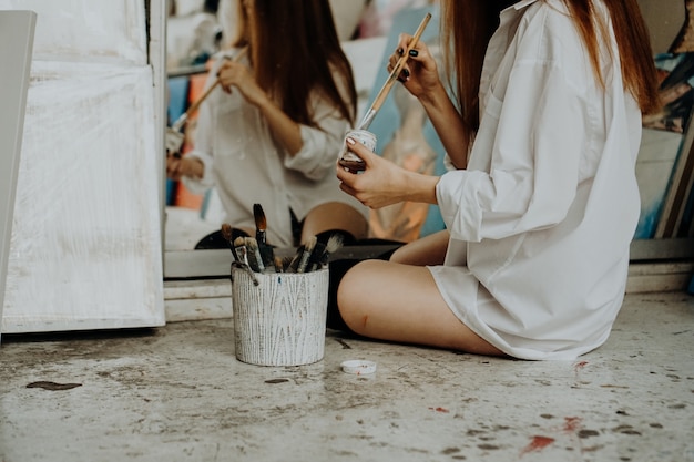 Pintor de la mujer sentada en el suelo delante del espejo y el dibujo. Interior del estudio de arte. Materiales de dibujo, pinturas al óleo, pinceles de artista