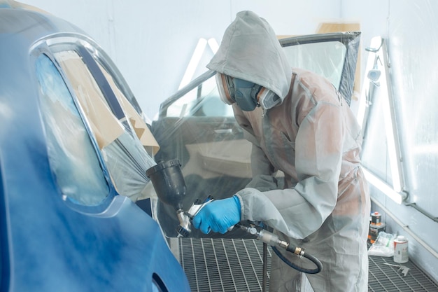 Pintor reparador de automóveis mão na luva protetora com aerógrafo pulverizador pintando o corpo do carro na câmara de pintura.