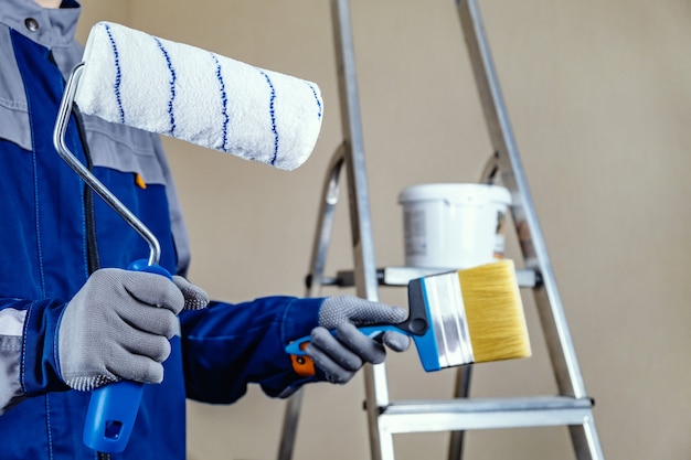 Foto el pintor de yeso está listo para pintar la pared. en manos de un rodillo y una brocha. una escalera de mano y un balde de pintura al fondo.