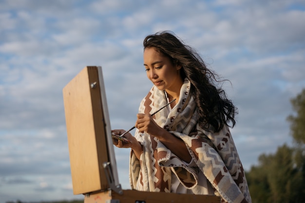 Pintora asiática en busca de inspiración, disfrutando de hermosos paisajes, sosteniendo pincel y paleta