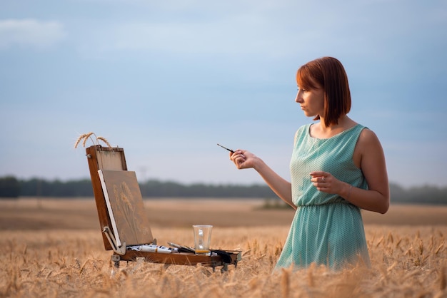 Pintora durante el trabajo al aire libre. Artista pelirroja haciendo las últimas pinceladas en su dibujo, complacida con el resultado de su trabajo.