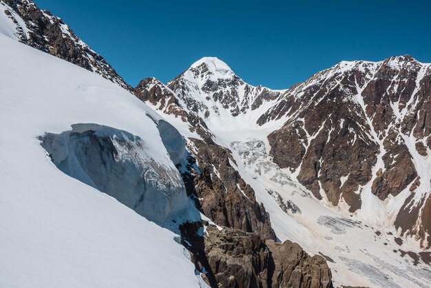 Pintoresco paisaje alpino con cima de montaña cubierta de nieve y lengua glaciar empinada con caída de hielo a la luz del sol Impresionante paisaje montañoso con pico puntiagudo y glaciar sobre rocas bajo un cielo azul en un día soleado