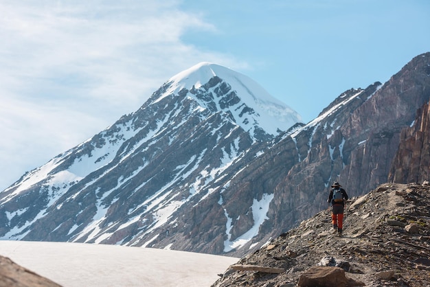 Pintoresco paisaje alpino con excursionista con bastones de trekking contra el pico de la montaña nevada y rocas afiladas a la luz del sol Hombre con mochila camina en altas montañas bajo cirros en el cielo azul en un día soleado